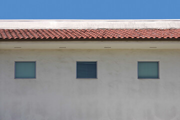 Fototapeta na wymiar Upper part of a white tile roof building under blue sky with three windows with closed blinds 