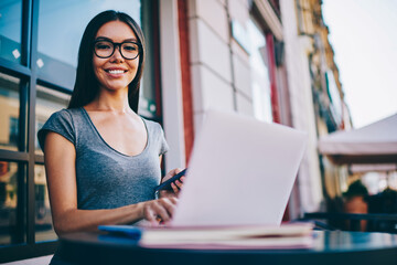 Portrait of cheerful female freelancer in eyewear satisfied with remote job in networks using application on phone, happy hipster girl share multimedia from smartphone synchronizing with laptop