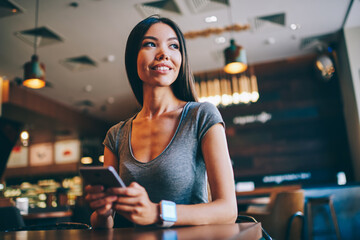 Dreamy positive female sitting with smartphone device in coffee shop, smiling female blogging in social networks share multimedia with followers using wireless connection and mobile phone gadget