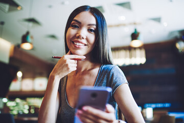 Portrait of happy caucasian hipster girl holding pen and smartphone gadget in hand and looking at camera during free time in cafeteria, smiling female blogger creating publication for content web page