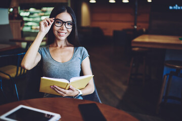 Portrait of happy hipster girl in optical spectacles feeling excited from time with favourite book in cafeteria, cheerful woman looking at camera while holding textbook for education indoors