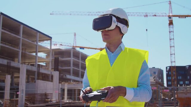 Professional drone operator in virtual reality helmet standing in front of construction site. Builder holding remote controller. Office building and crane background.