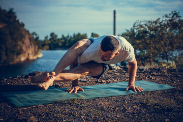 Fit muscular man doing yoga by the water on the grass with hands stretch. Orange sunlight at quarry lake. Healthy spine fitness workout. International Day of Yoga