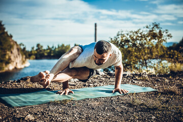 Fit muscular man doing yoga by the water on the grass with hands stretch. Orange sunlight at quarry lake. Healthy spine fitness workout. International Day of Yoga