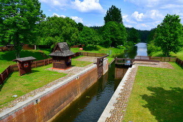 Fototapeta na wymiar View of a massive dam made of wood and bricks with a small hut for monitoring the condition of the object located nearby, next to a dense forest or moor seen on a cloudy yet warm summer day in Poland