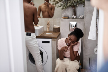 morning of young african married couple, they brush teeth at one time, woman sits on toilet bowl while her husband looks at mirror - Powered by Adobe