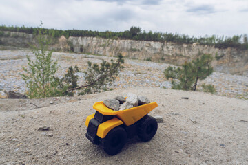 Small yellow car toy, mining truck with stones at the hill at the background of quarry, toned, copy space. Stones and trees at background and blue sky.