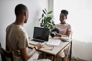 young black woman with camera in hands talk with husband working on laptop, share opinions, work at home together