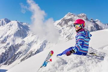 Happy young girl in bright sport outfit sit on the snow pile in the mountain over high peaks look back to camera