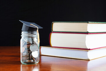 Graduation cap on the  glass jar with coins and in front of books stack.Concept of saving for education.