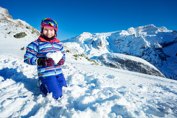 Fototapeta na wymiar Ski little girl hold snow in heart shape smiling with mountain summit on background standing on her knees
