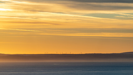 Wind farm during very golden hour with wind turbines at the cliff top at the horizon above calm sea under golden morning sky