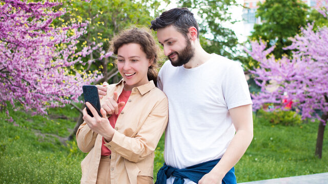 Smiling young family couple using smartphone,standing in blooming city park.Happy brunette man and woman finger pointing on mobile phone screen,wireless internet connection,shopping online,technology