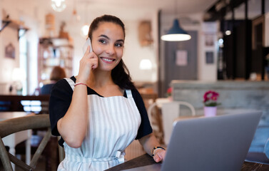 Young woman with apron, smartphone and laptop working in cafe.