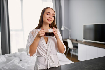 attractive young caucasian lady is enjoying smell of hot coffee, woman inhales aroma of coffee in bedroom