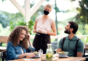Fototapeta premium Waitress with face mask serving happy couple outdoors on terrace restaurant.