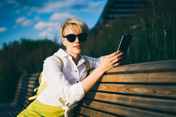 Half length portrait of stylish young woman with short haircut and trendy black sunglasses looking at camera while sitting on wooden bench with modern telephone in hand outdoors in summer weather