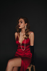 Woman with wavy hair dressed in red silk outfit posing on black background. Elegant lady in satin dress sitting on isolated backdrop