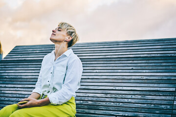 Young woman enjoying favourite music in earphones connected to telephone sitting on wooden bench in...