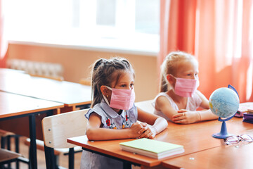 Fototapeta na wymiar Two little schoolgirls sit at a desk in a school class in medical masks and carefully listen to the teacher