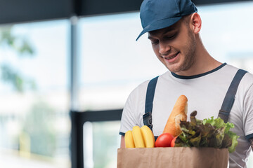 Fototapeta premium cheerful delivery man holding paper bag with fresh groceries
