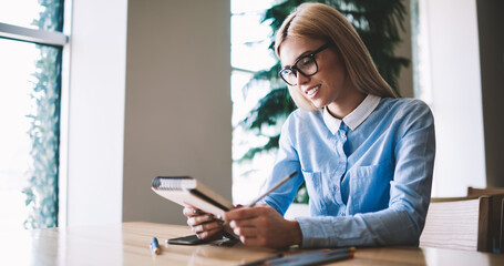 Happy talented female illustrator for child books looking on pictures draw in textbook for education, positive woman student enjoying learning indoors while reading information from notebook