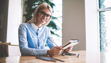 Portrait of cheerful blonde hipster girl learning in university class room with loft interior, happy smiling female student in eyewear holding knowledge textbook in hands and looking at camera