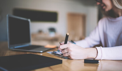 Selective focus on female hand writing information via elegant pen while using modern laptop computer for making research, woman messaging via netbook application on blurred background