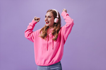 Charming lady in pink hoodie dancing on purple background and enjoying songs in headphones. Portrait of happy blonde woman in sweatshirt listening to music