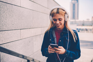 Smiling young woman in denim wear choosing music on internet website to download in playlist on phone.Positive student listening songs in headphones connected to cellular while chatting online