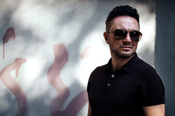 stylish guy with glasses looks aside against the background of a spotted fence. strong and strict male gaze. brown T-shirt with collar. loose sunlight. chest portrait