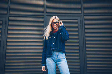 Below portrait of stylish positive young woman with blonde hair posing at camera.Cheerful hipster girl in optical eyeglasses dressed in denim clothes standing outdoors on black promotional background