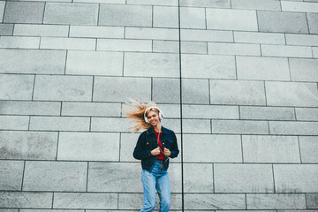 Obraz na płótnie Canvas Cheerful blonde young woman in stylish wear listening audio music in modern headphones jumping outdoors against grey wall.Carefree hipster girl dressed in denim apparel enjoying electronic songs