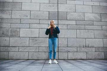 Obraz na płótnie Canvas Stylish young woman dressed in trendy denim clothes standing outdoors on grey promotional background with copy space while listening favourite music from playlist in modern white headphones