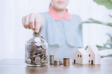 Half face of Asian woman that hand holding coin act likes put coin into jar glass bank next to stacking coins and house wood model. Money saving and house loan financial concept.