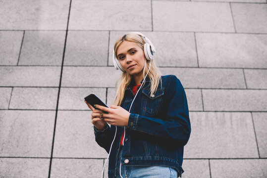 Below portrait of blonde attractive female blogger listening audio songs in modern headphones while installing mobile application with new music on smartphone via 4G internet standing near grey wall