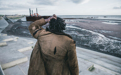A black male with dreadlocks and a ripped tatty homeless style coat and clothes looking over a...