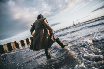 Naklejka premium A black male with dreadlocks and a ripped tatty homeless style coat and clothes looking over a dirty cold post apocalyptic nuclear war water sea setting. Conceptual future earth .