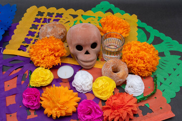 Mexican Day of the dead altar with traditional bread, colorful flowers, a skull, mezcal, candles and cut paper on black background
