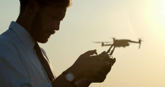 Side view of bearded man in white shirt and black tie holding remote controller while drone is flying over green field. Professional agronomist scanning harvest during amazing sunset.