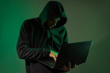 A young man in a hood with a laptop in his hands. A hacker makes a hack through a netbook