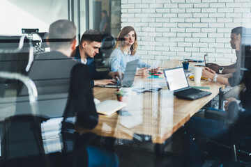 Team of multicultural male and female professionals dressed in formal wear discussing productive strategy together with proud ceo sitting at meeting table in stylish office interior behind glass wall