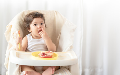 Obraz na płótnie Canvas Messy baby girl eating cake. Lovely infant girl holding a spoon, enjoying eating cake.