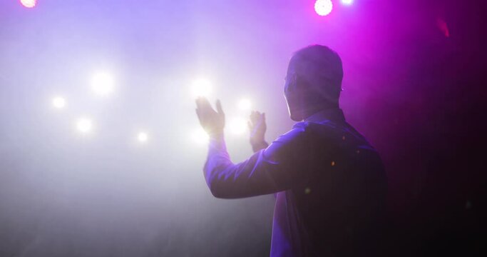 Showman stand up comedian is clapping hands on stage and entertaining audience at the end of the performance or show in spotlights and white theatral smoke on light background, side view.