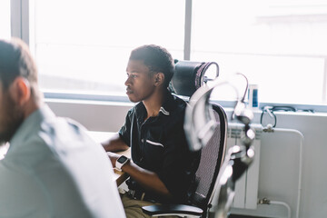 Pensive african american young man dressed in formal wear sitting at meeting table having brainstorming session in office.Dark skinned attentive employee listening colleague during briefing