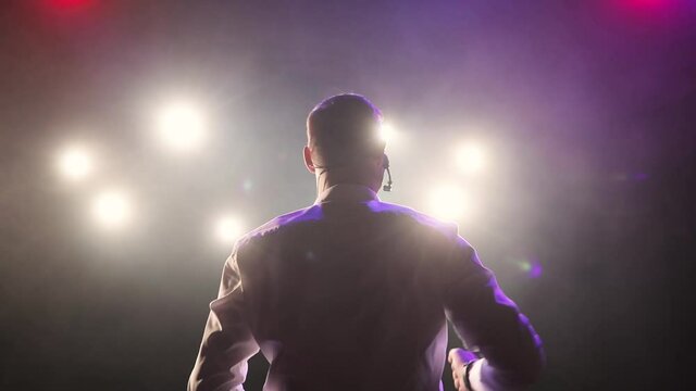 Man stand up comedian speaking jokes in micropphone in spotlights standing on stage with his back to the camera. Actor on funny performance, concert, show. Actively gestures with his hands.