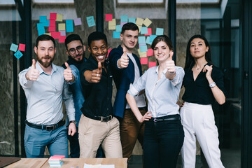 Portrait of multicultural team of happy office employees dressed in formal wear holding thumb up showing sign okay and success of collaboration standing against wall with colorful stickers in office