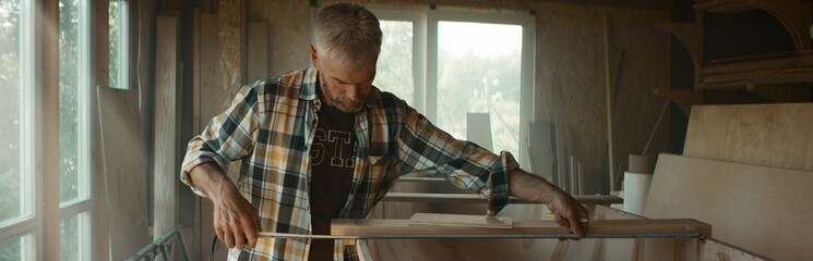 Caucasian male using measure tape while building a canoe in his workshop. Boat making hobby, small...