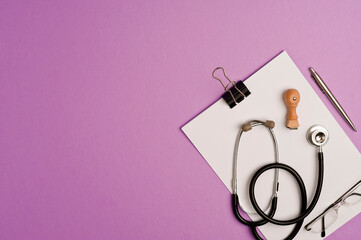 Medicine accessories concept, phonendoscope on top of notepad with silver pen, eyeglasses, ok vaccine stamp, lilac background with copy space. top view