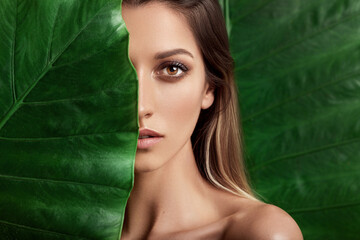 Luxury Portrait of a Beautiful young woman with natural makeup holds a big green leaf on a blurred green background. Spa and wellness. Youth, teens and skin care concept. Close up